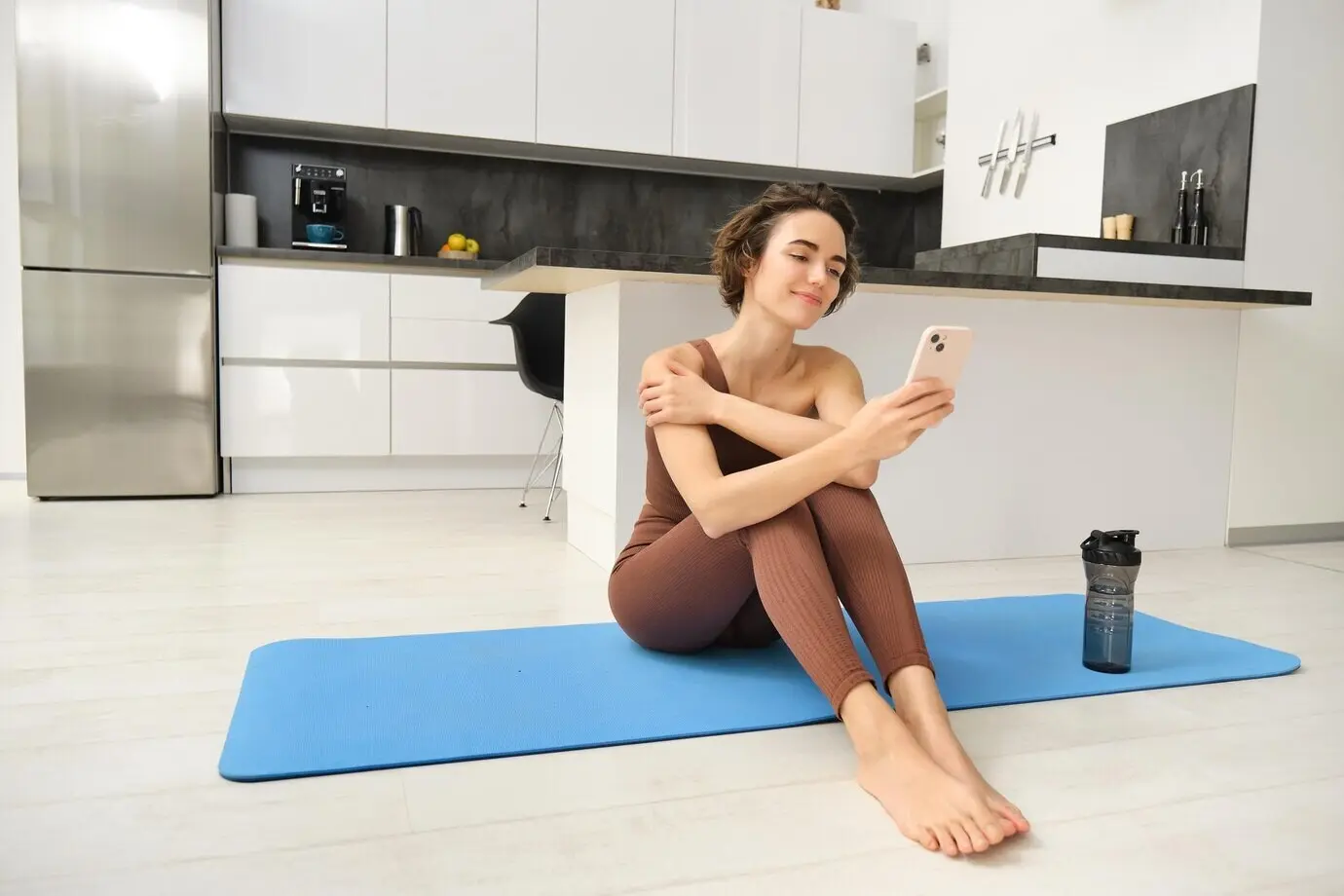 Porträt eines sportlichen Mädchens mit Mobiltelefon, das auf einer Yogamatte sitzt und während des Workouts zu Hause ein Smartphone benutzt.